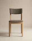 The Wooden Stories Noki Dining Chair in Light Oak features contract grade solid wood, a cushioned seat and back in textured gray fabric, and is shown against a beige background.