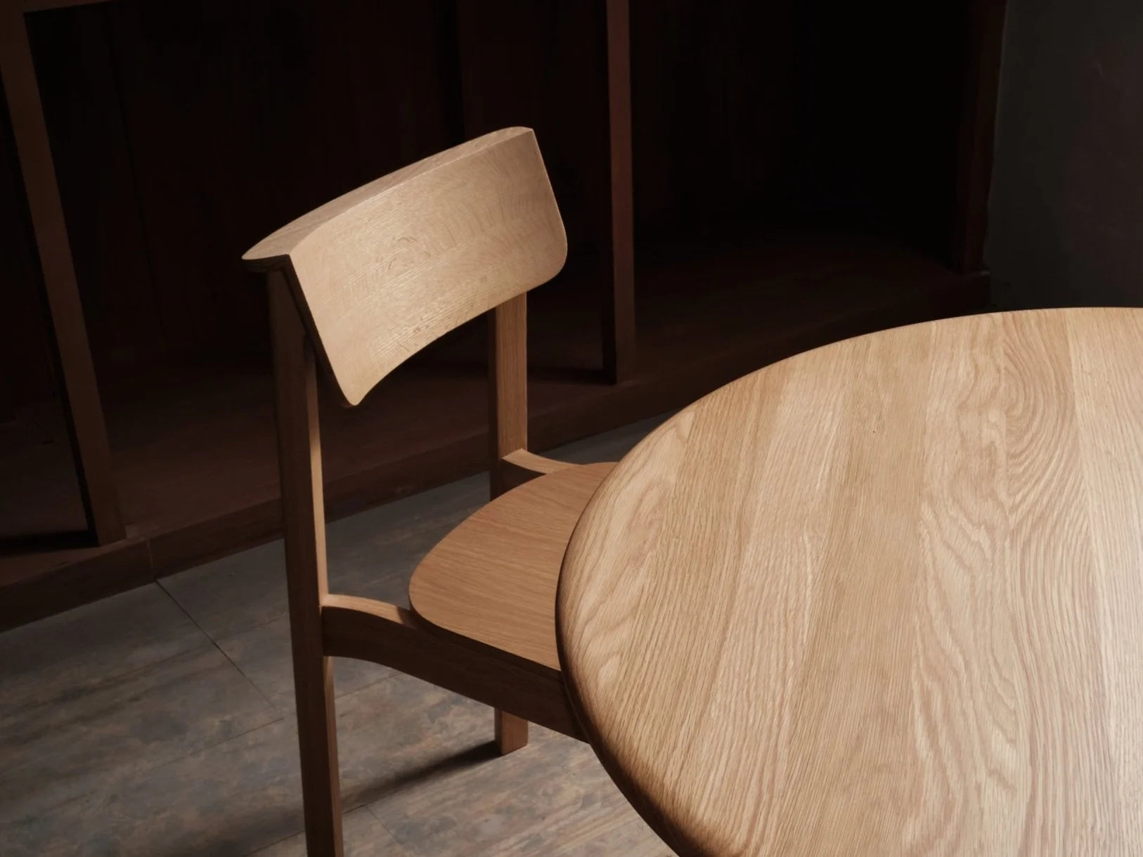 A Noki Dining Chair in Light Oak by Wooden Stories, featuring a curved backrest and natural finish, sits next to a round wooden table. The contract-grade setting is indoors with soft lighting.