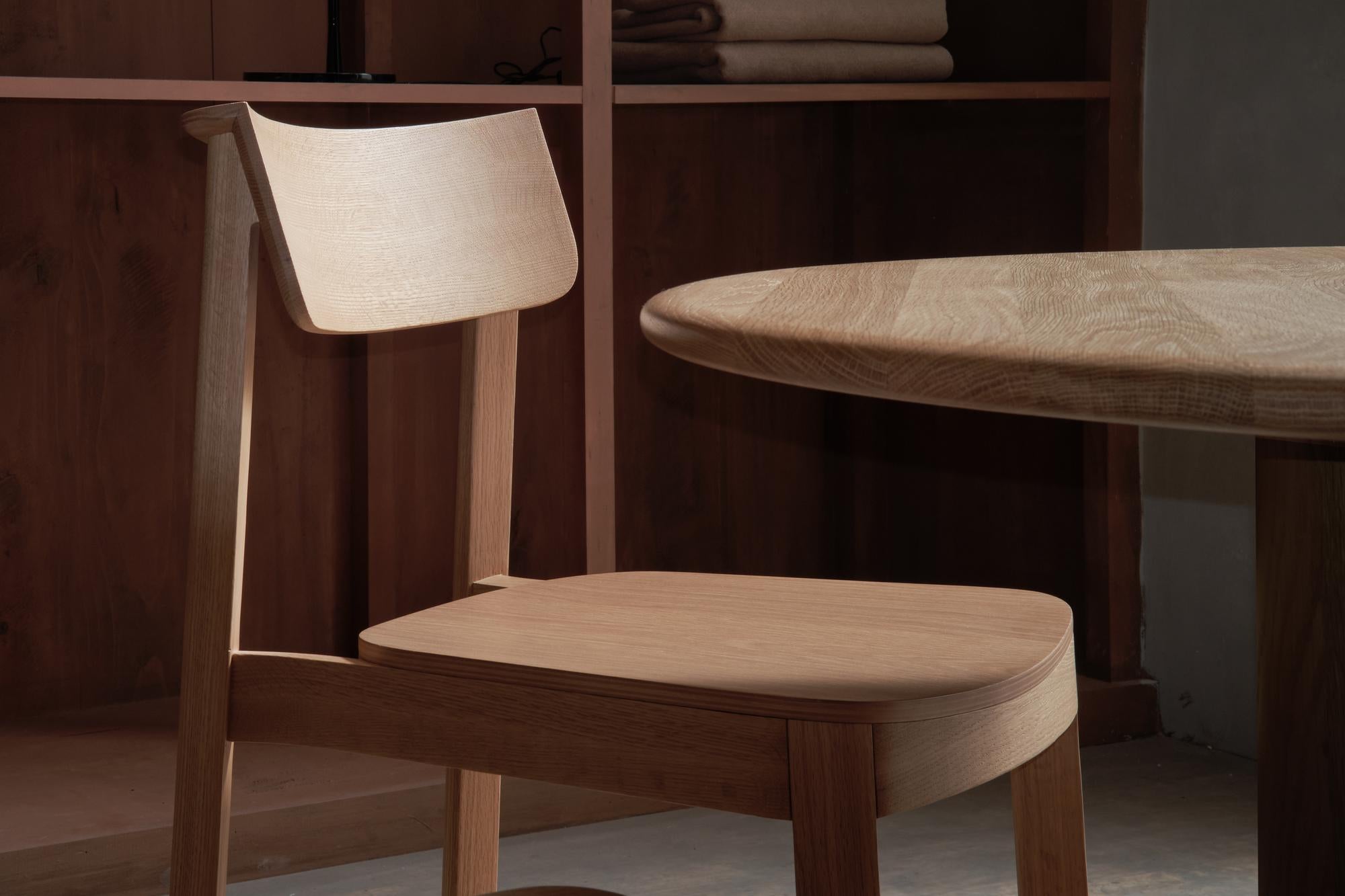 A Wooden Stories Noki Dining Chair | Light Oak with a curved backrest sits by a round wooden table in a warmly lit room, accented by shelves and folded blankets in the background.