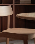 A Wooden Stories Noki Dining Chair | Light Oak with a curved backrest sits by a round wooden table in a warmly lit room, accented by shelves and folded blankets in the background.