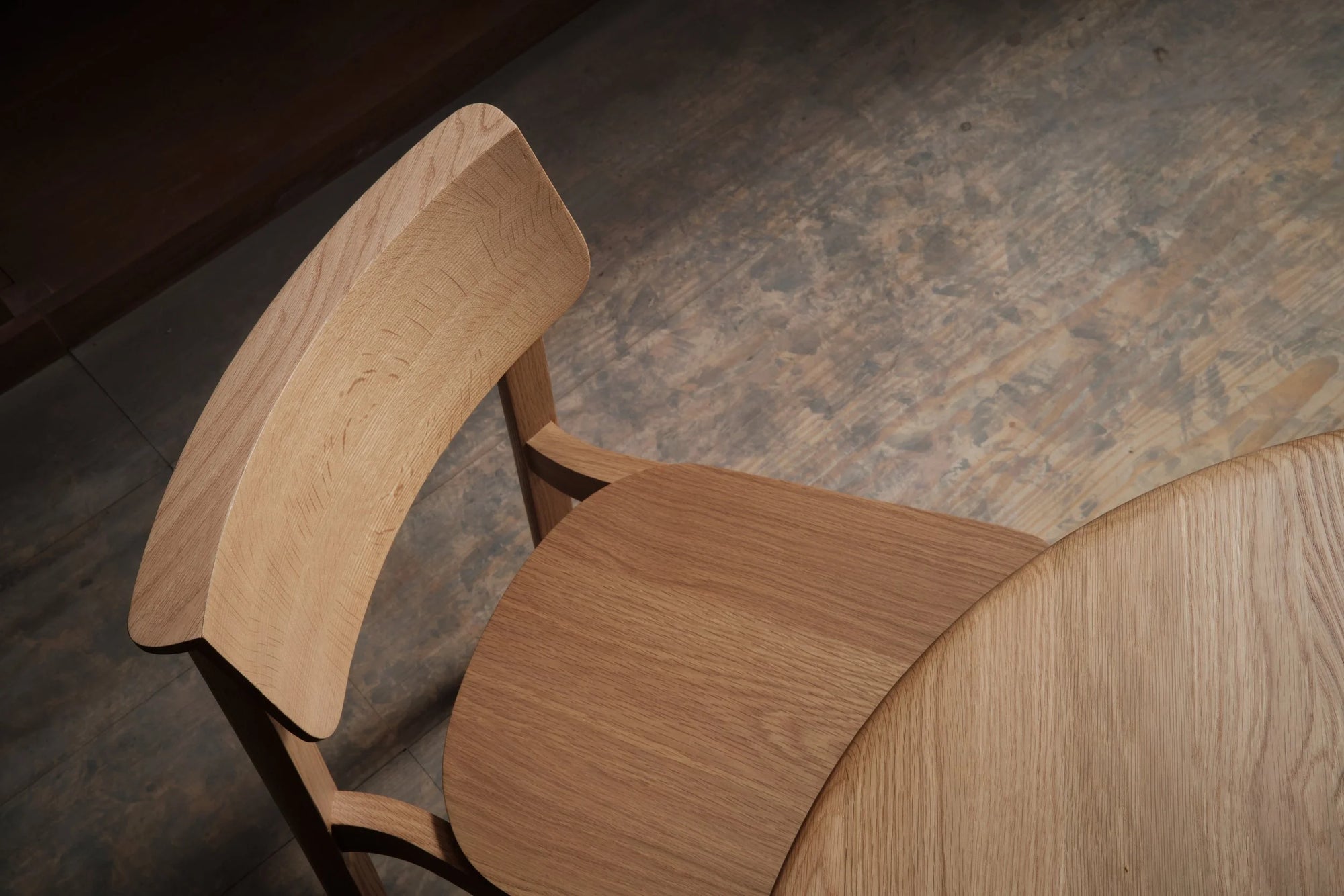 The Noki Dining Chair | Light Oak by Wooden Stories, featuring a curved backrest and natural wood finish, sits beside a round wooden table on a textured floor. The contract grade chair is photographed from above.