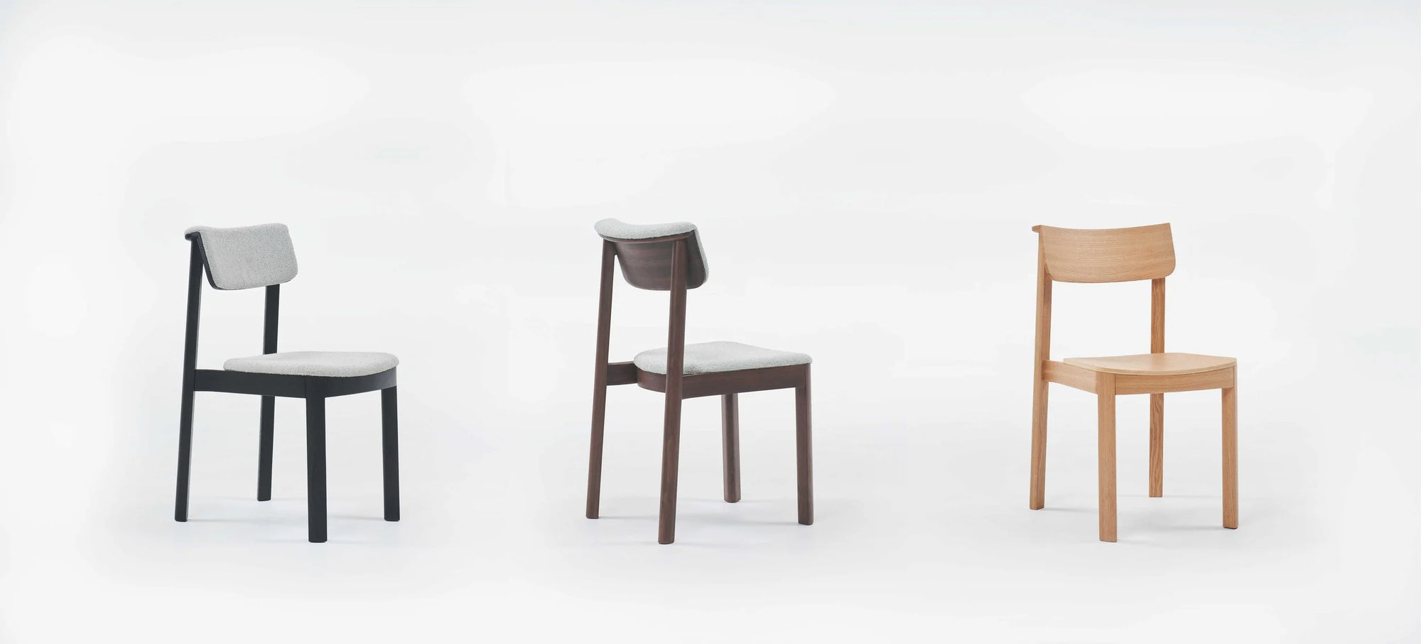 Three Wooden Stories Noki Dining Chairs in Espresso Oak are lined up against a plain white background. Each contract-grade solid wood chair features a unique color: black with gray fabric, dark brown with gray fabric, and light wood without upholstery.