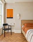 Minimalist bedroom featuring the Ethnicraft Nordic II Bed, crafted from European oak and dressed in orange bedding with a striped blanket. The room includes a black chair, a compact desk, books, wall art in orange and black tones, and a narrow window that lets natural light fill the space.