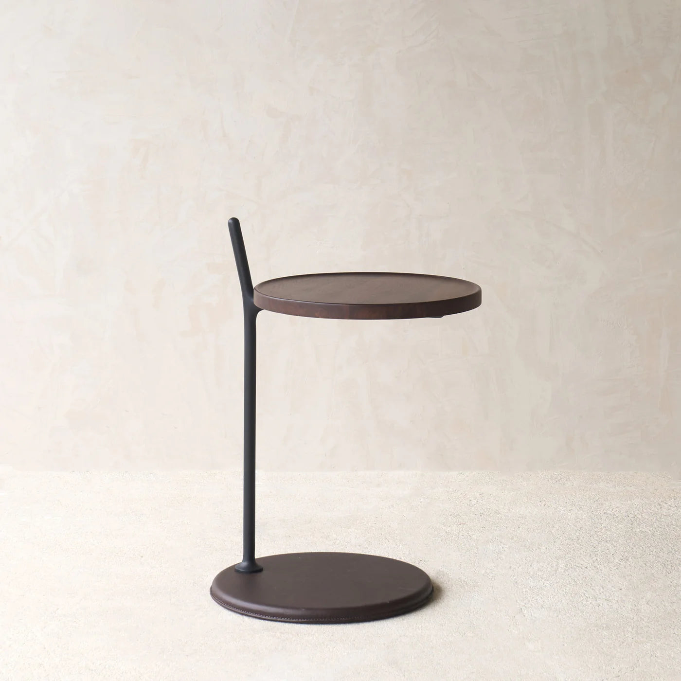 The Bystander Side Table by Wooden Stories features a round dark brown top and base, joined by an offset slim black support, shown on a light textured surface against a beige wall.
