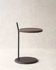 The Bystander Side Table by Wooden Stories features a round dark brown top and base, joined by an offset slim black support, shown on a light textured surface against a beige wall.