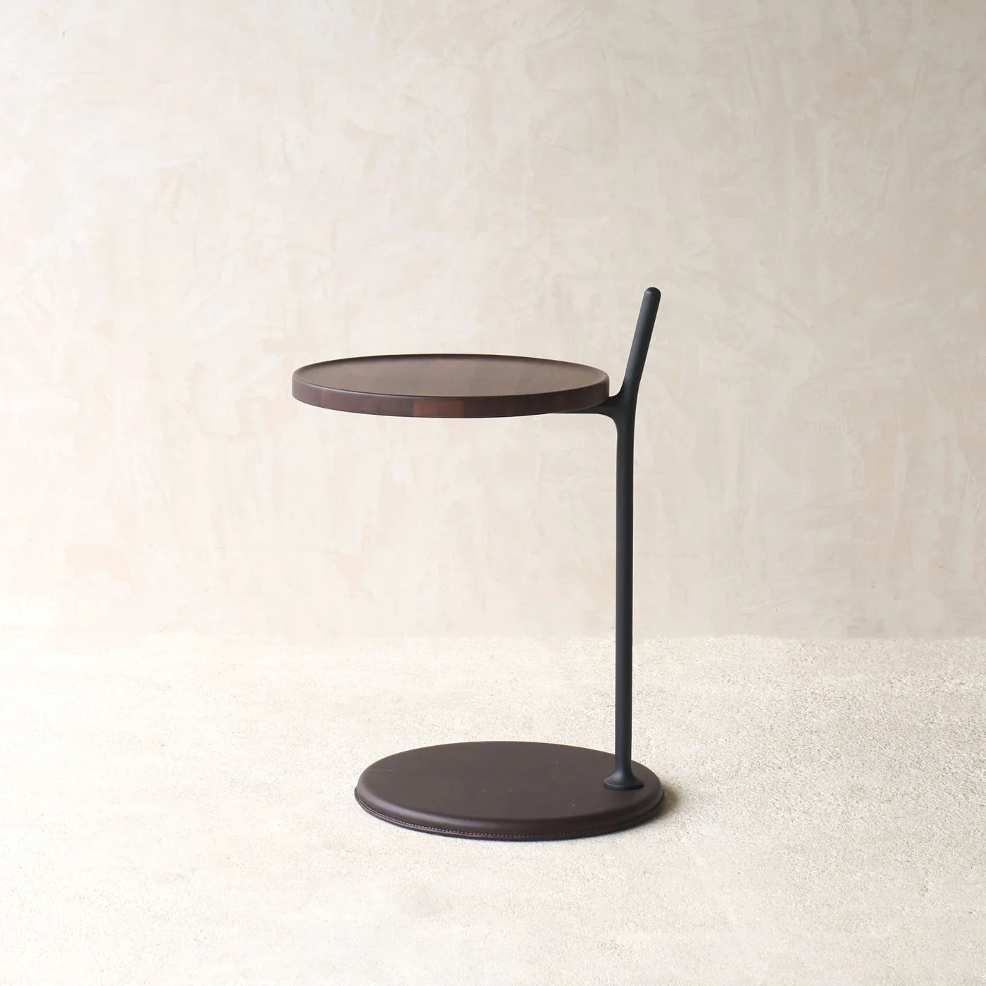 The Bystander Side Table by Wooden Stories features a modern minimalist design with a round dark wood top and base, connected by a single black vertical support, shown against a light beige textured wall and neutral floor.