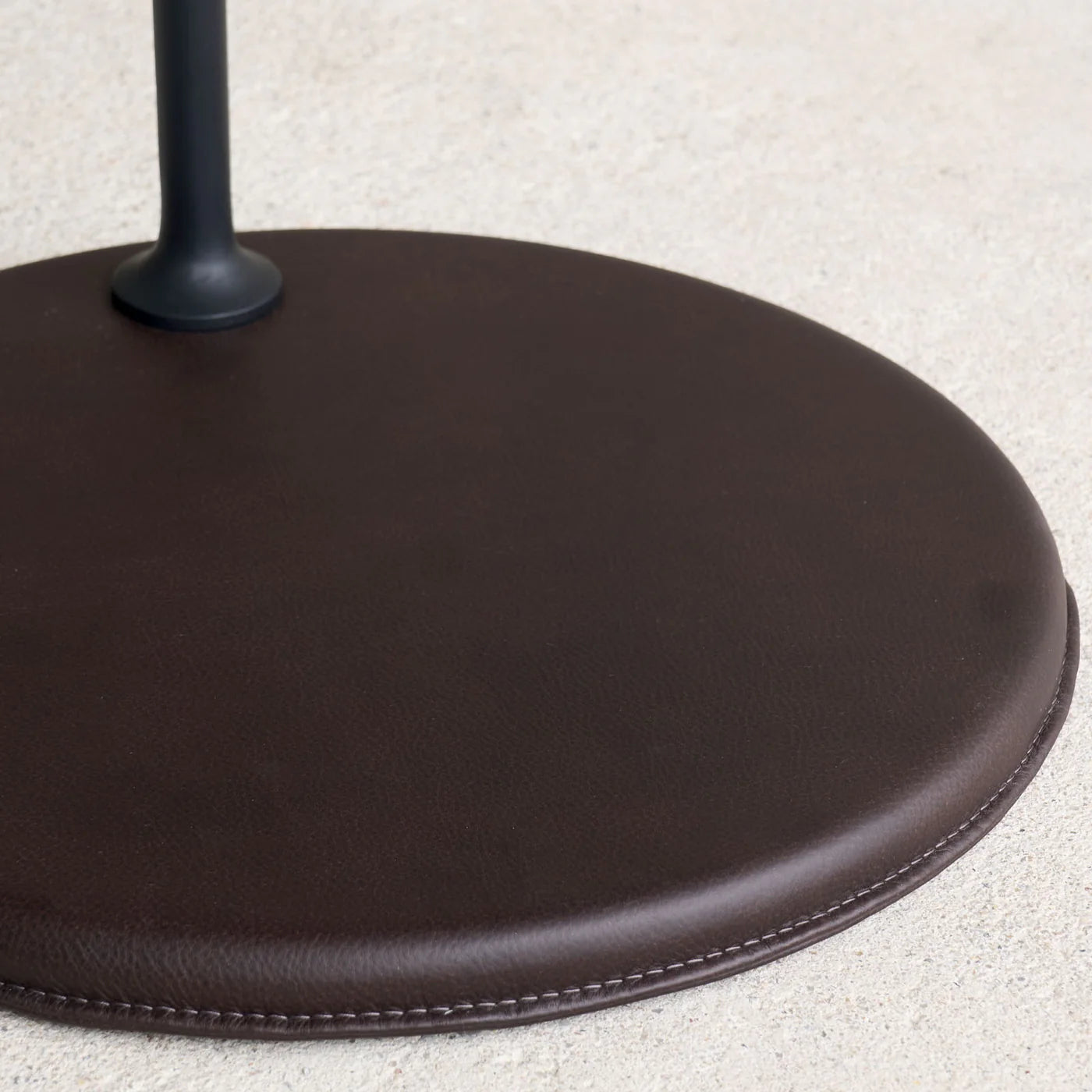 A close-up of the Wooden Stories Bystander Side Table shows its round, dark brown leather base with visible stitching on a light-textured surface and a black metal pole attached at the top left.