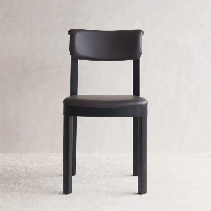 The Noki Dining Chair by Wooden Stories features black oak and a padded brown leather seat with a curved backrest, inspired by Japanese design, placed on a light carpet against a beige wall.