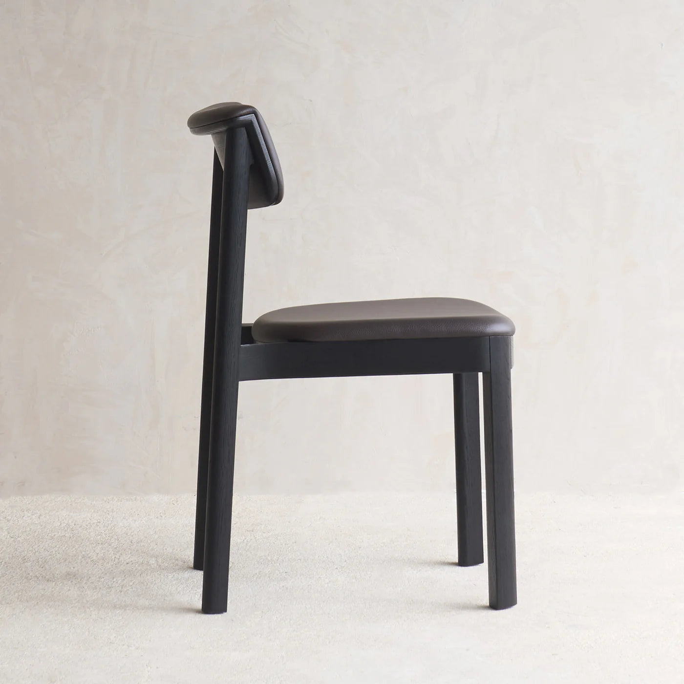 The Noki Dining Chair by Wooden Stories, in black oak with a leather seat and backrest inspired by Japanese design, is shown in profile on a light textured floor against a neutral beige wall.