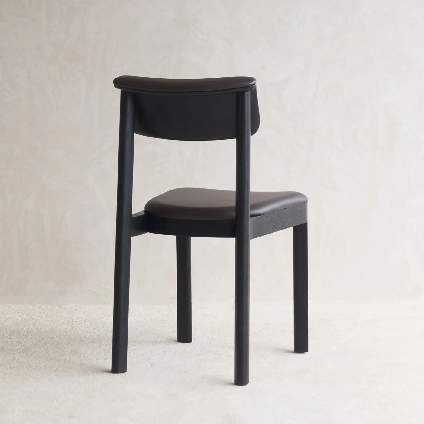 The Noki Dining Chair - Black Oak - Leather by Wooden Stories, featuring a minimalist Japanese-inspired design with a curved back and padded seat, is set on a light textured floor against a neutral wall, facing away from the camera.