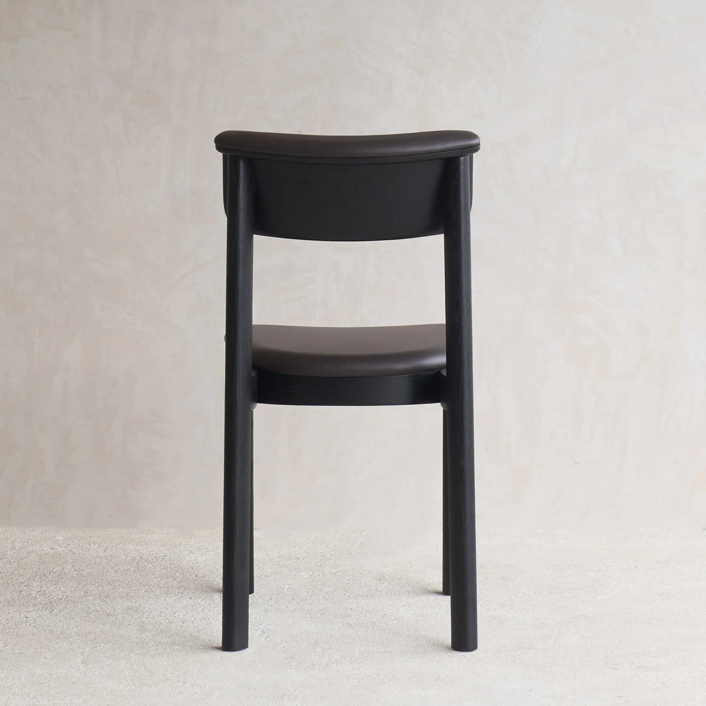 The Noki Dining Chair by Wooden Stories, in Black Oak and Leather, designed by Ryota Yokozeki with Japanese inspiration, features a curved backrest and seat. Shown from behind on a light textured floor against a neutral wall.