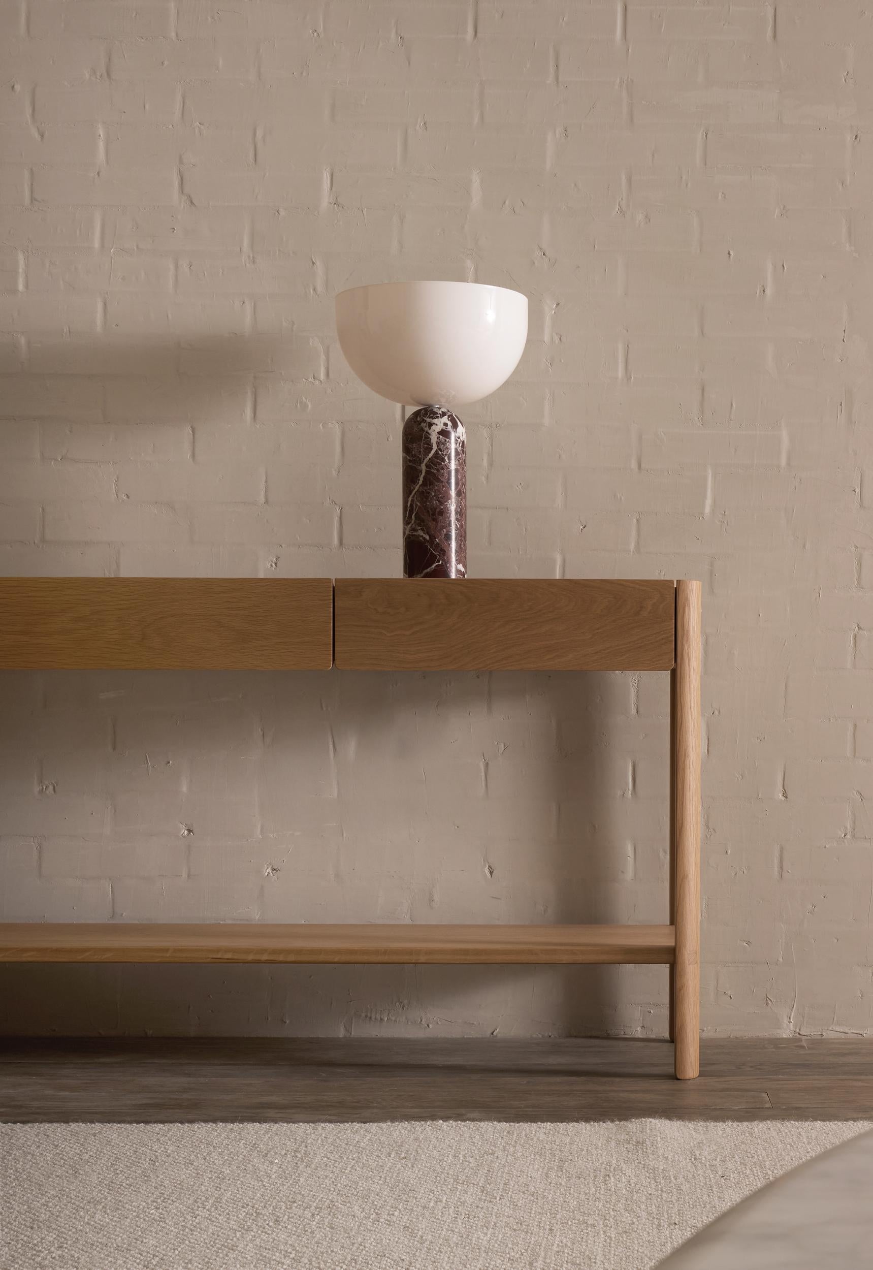 The Wooden Stories Nell Console in Light Oak, inspired by Danish design, stands against a light beige brick wall, topped with a modern lamp with a white bowl shade and dark marble base. A light-colored rug is visible in the foreground.