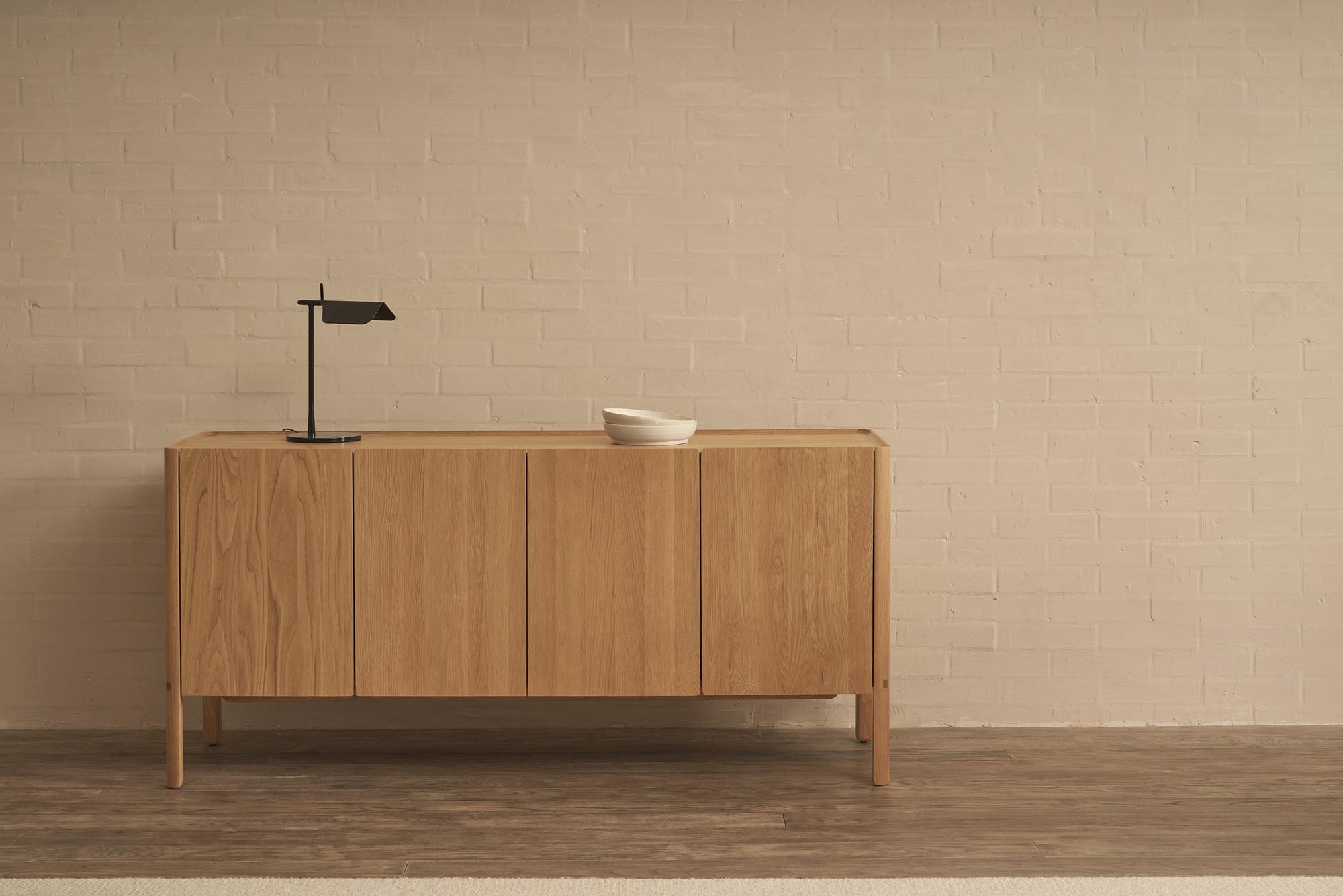 The Nell Sideboard | Light Oak by Wooden Stories, with spacious storage, stands against a light beige brick wall. On top are a small black lamp and two white bowls, while the floor combines wood and a light-colored carpet.