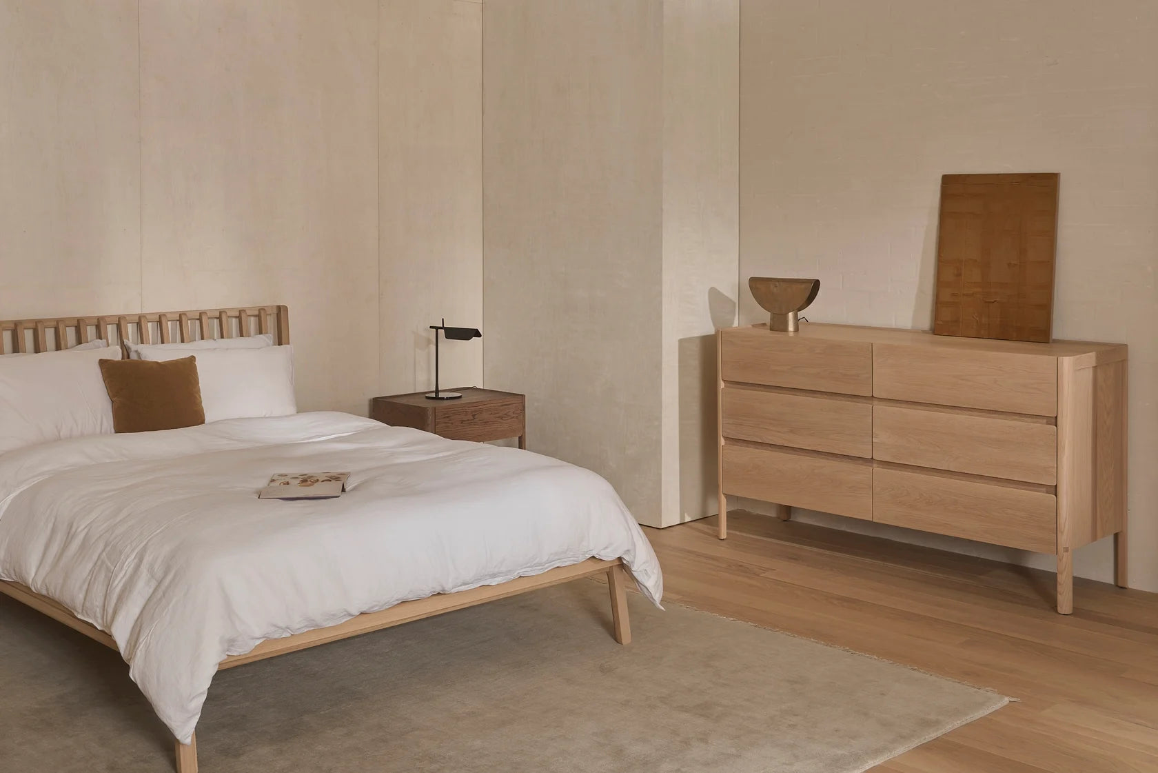Minimalist bedroom featuring a wooden bed frame, crisp white bedding, a brown accent pillow, wooden nightstand, black lamp, and the Nell Tall Dresser by Wooden Stories with premium metal runners for smooth bedroom storage solutions. Decor accents and a beige rug on hardwood floors complement the neutral tones and simple, modern design inspired by the Danish Collection.