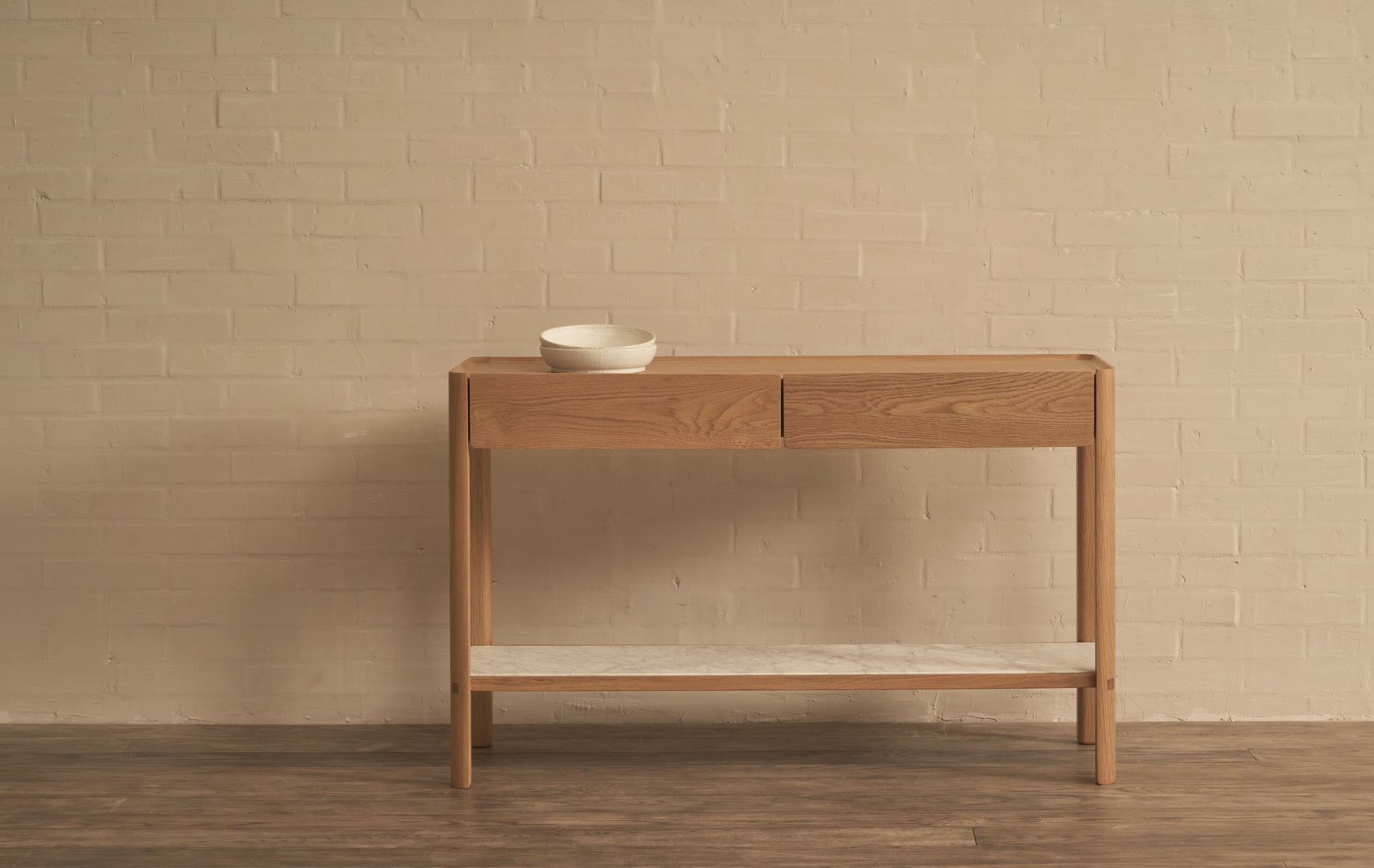 The Nell Console by Wooden Stories, in light oak with stone, features solid oak construction, two drawers, and a white lower shelf. Set against a beige brick wall on a dark wood floor, it’s topped with two white ceramic bowls.