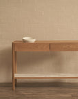 The Nell Console by Wooden Stories, in light oak with stone, features solid oak construction, two drawers, and a white lower shelf. Set against a beige brick wall on a dark wood floor, it’s topped with two white ceramic bowls.
