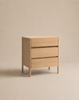 The Nell Tall Dresser by Wooden Stories, featuring a minimalist light wood design and three spacious drawers with premium metal runners for smooth operation, stands elegantly on four legs against a plain beige background, offering refined bedroom storage.