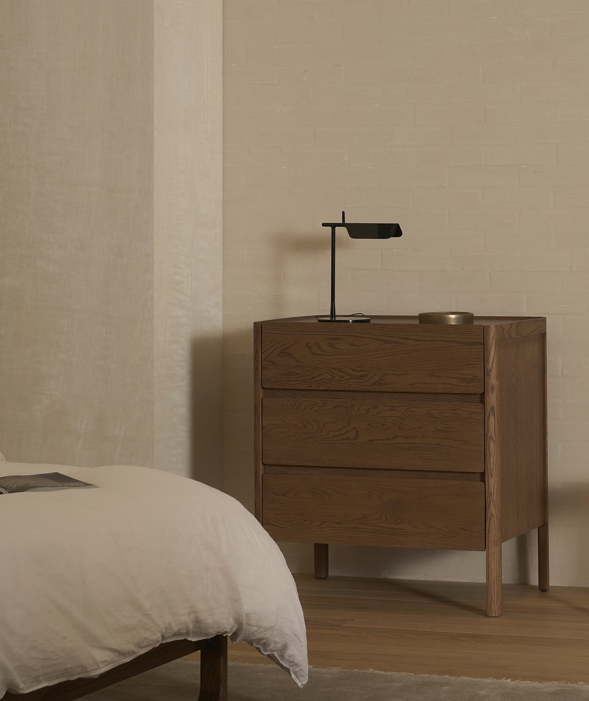 A minimalist bedroom showcases the Nell Tall Dresser by Wooden Stories, featuring premium metal runners, paired with a black desk lamp and a round tray. Part of a bed with white bedding appears in the foreground. The room provides ample bedroom storage and highlights the calm, neutral aesthetic typical of the Danish Collection.