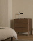 A minimalist bedroom showcases the Nell Tall Dresser by Wooden Stories, featuring premium metal runners, paired with a black desk lamp and a round tray. Part of a bed with white bedding appears in the foreground. The room provides ample bedroom storage and highlights the calm, neutral aesthetic typical of the Danish Collection.