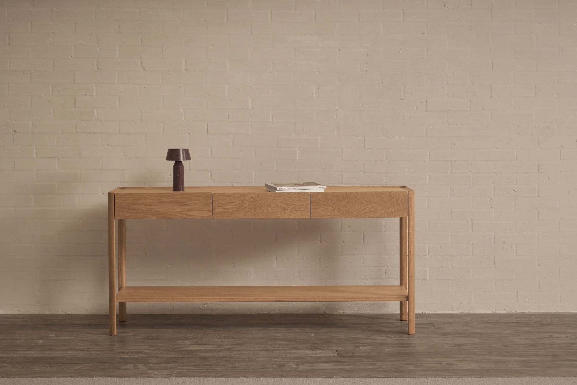 The Nell Console in Light Oak by Wooden Stories, featuring three drawers and a lower shelf, stands against a beige brick wall. A small purple lamp and closed book rest on top, complementing the dark wood floor.