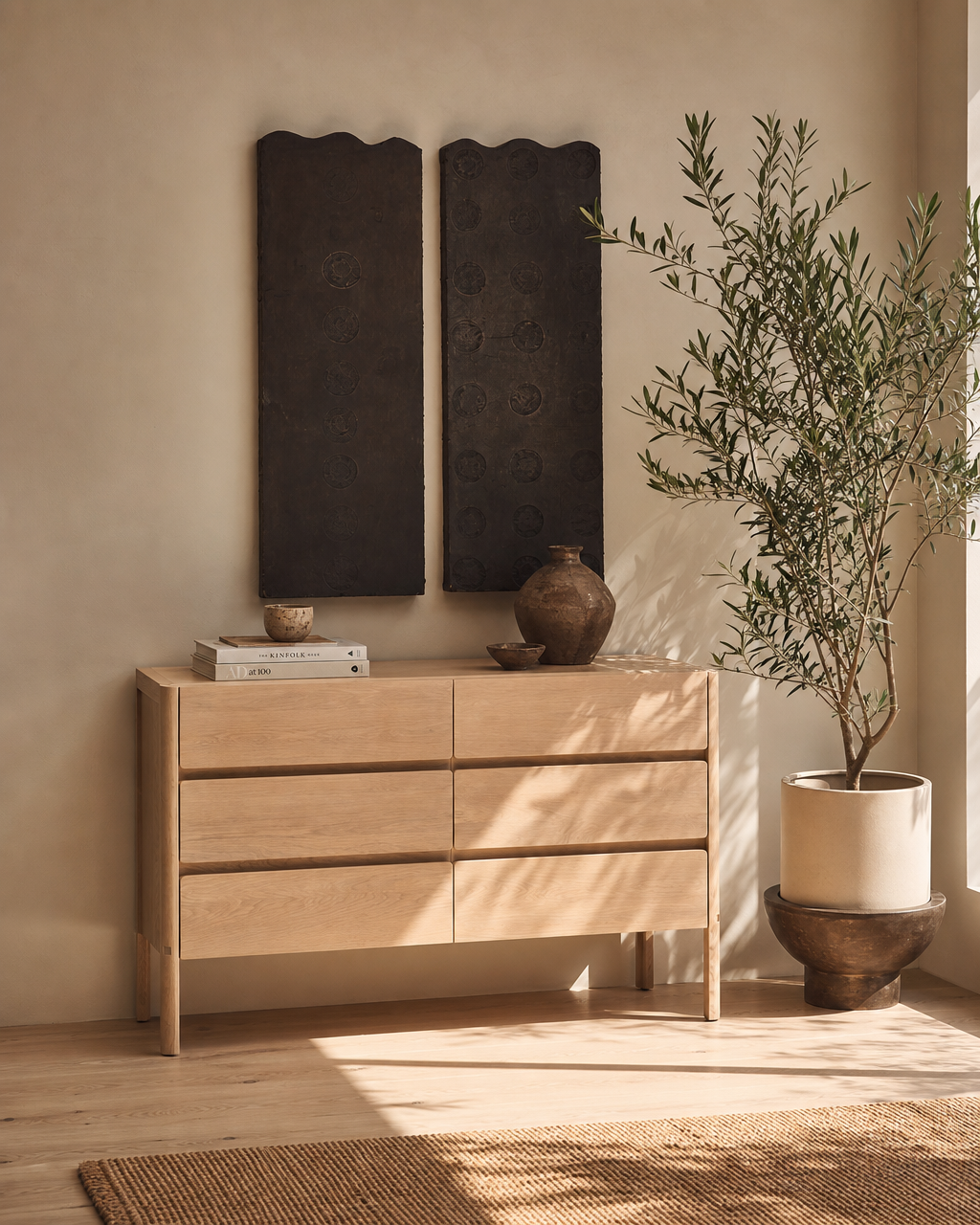 The Nell Tall Dresser by Wooden Stories, crafted in light wood with premium metal runners and accents, pairs beautifully with two dark wall panels, a potted tree, and sunlight streaming in to create a serene minimalist interior of neutral tones—an elegant expression of the Danish Collection approach to bedroom storage.