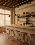 A minimalist bar featuring Wooden Stories' Odd Counter Stool | Leather: Italian leather upholstered seats and wooden bases line a marble counter, with pendant lights overhead, shelves of bottles on a white brick wall, and large windows offering natural light.
