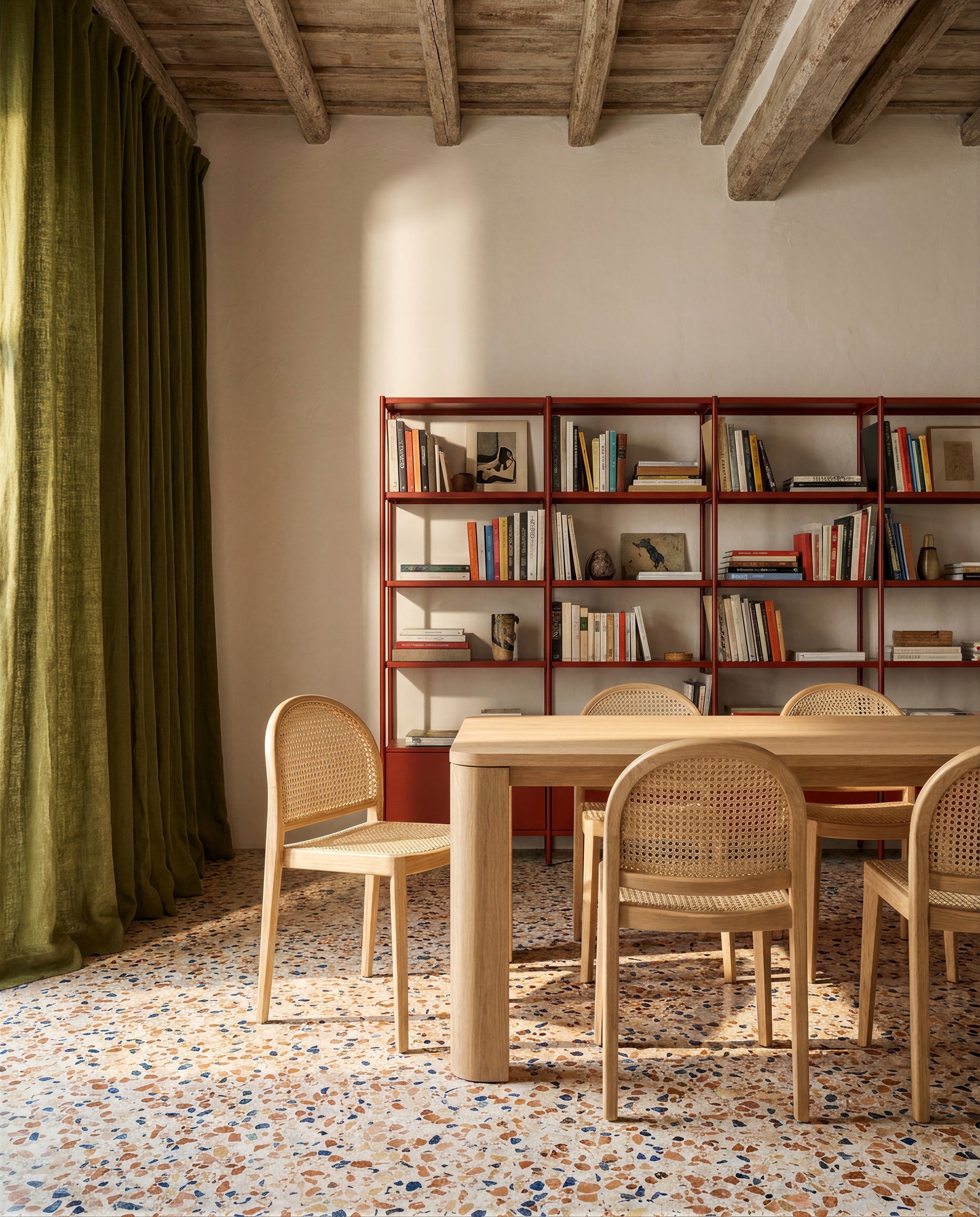 A sunlit room with a terrazzo floor features the Oli Dining Table by Wooden Stories and cane chairs. A red metal bookshelf against the wall displays books and decor, while green curtains frame the window and wooden beams cross the ceiling.