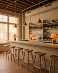 A minimalist bar with Danish design, a light wooden counter, six Wooden Stories Odd Counter Stools in light oak with leather seats, pendant lights, bottle-lined shelves, exposed brick walls, large windows, and warm ambient lighting.
