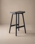 The Odd Counter Stool | Wood by Wooden Stories, a modern black solid oak stool with a curved seat and four angled legs, displays smooth edges and minimalist Danish design against a neutral beige background.