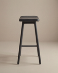 The Odd Counter Stool | Leather by Wooden Stories features a minimalist design with four legs and a square leather seat, ideal for kitchen island seating, shown against a light beige background.