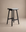 The Odd Counter Stool | Fabric by Wooden Stories showcases Danish minimalist design with a cushioned gray seat and black wooden legs, crafted from natural materials, displayed on a plain beige background.