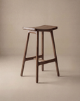 The Odd Counter Stool | Wood by Wooden Stories features a solid oak build, curved seat, and four angled legs with horizontal supports—ideal for your kitchen island, shown here against a plain beige background.