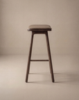 The Odd Counter Stool | Leather by Wooden Stories features a minimalist design with a rectangular seat and four angled legs, showcased against a plain beige background.
