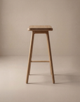 The Odd Counter Stool by Wooden Stories features a solid wood design with four legs and a rectangular seat, complementing minimalist, neutral-toned spaces with soft lighting.
