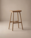 The Odd Counter Stool | Leather by Wooden Stories features a minimalist, modern design with a curved seat, four straight legs, and a natural wood finish—ideal as kitchen island seating and shown against a neutral beige background.