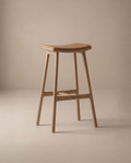The Odd Counter Stool | Leather by Wooden Stories features a minimalist, modern design with a curved seat, four straight legs, and a natural wood finish—ideal as kitchen island seating and shown against a neutral beige background.