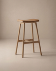 The Odd Counter Stool | Leather by Wooden Stories features a minimalist, modern design with a curved seat, four straight legs, and a natural wood finish—ideal as kitchen island seating and shown against a neutral beige background.