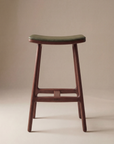 The Odd Counter Stool | Leather by Wooden Stories features four solid oak legs, a curved seat, and green Italian leather upholstery, shown on a neutral beige background.