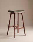 The Odd Counter Stool | Leather by Wooden Stories features a minimalist solid oak frame, dark green curved leather seat, and four angled legs—ideal for stylish kitchen island seating against a neutral beige backdrop.