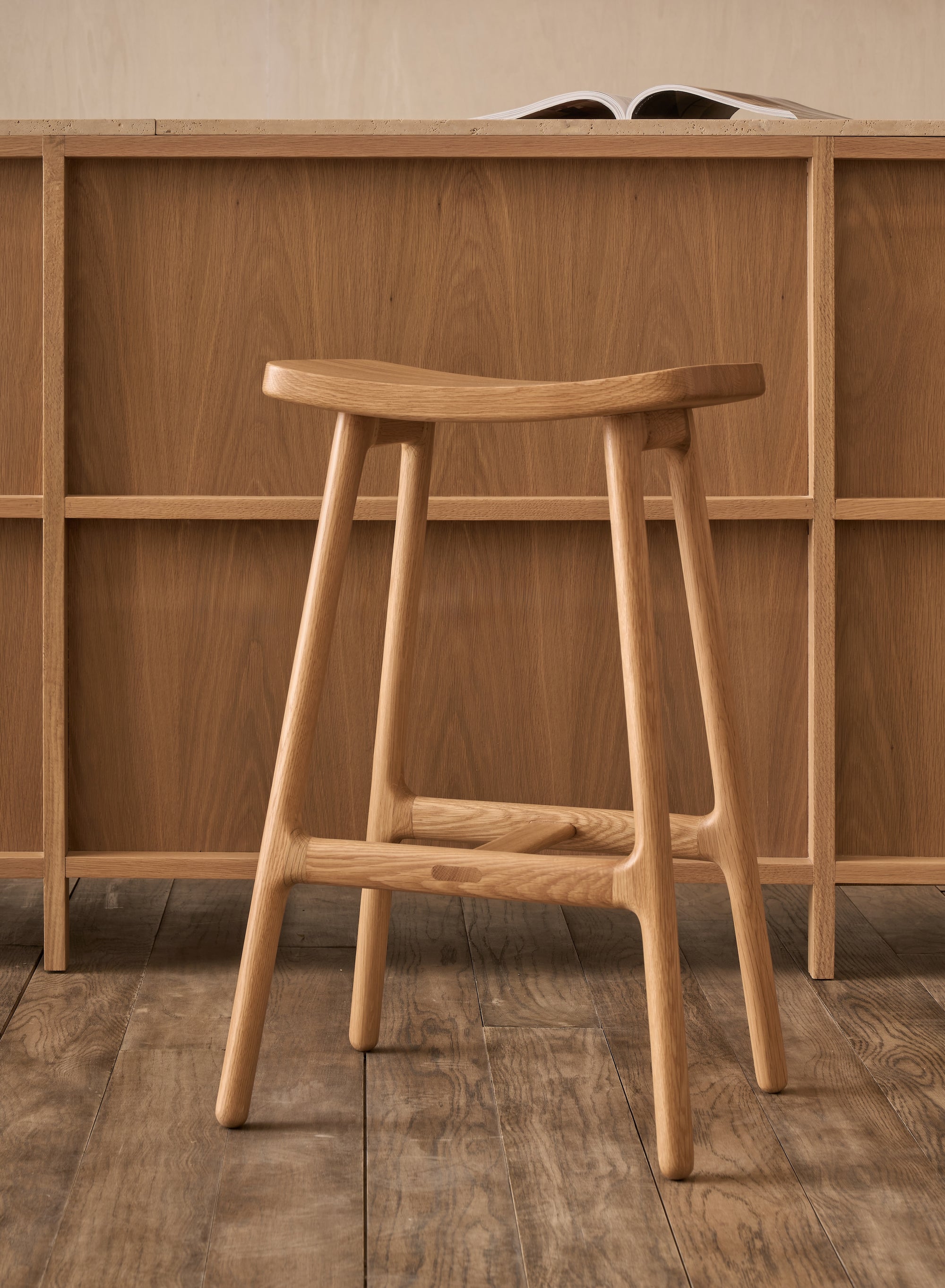 The Odd Counter Stool | Wood by Wooden Stories, featuring a curved solid oak seat in a light finish, is paired with a matching desk. An open book sits atop, while the visible wood grain below reflects Danish design influence.