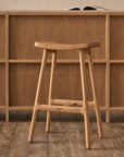 The Odd Counter Stool | Wood by Wooden Stories, featuring a curved solid oak seat in a light finish, is paired with a matching desk. An open book sits atop, while the visible wood grain below reflects Danish design influence.
