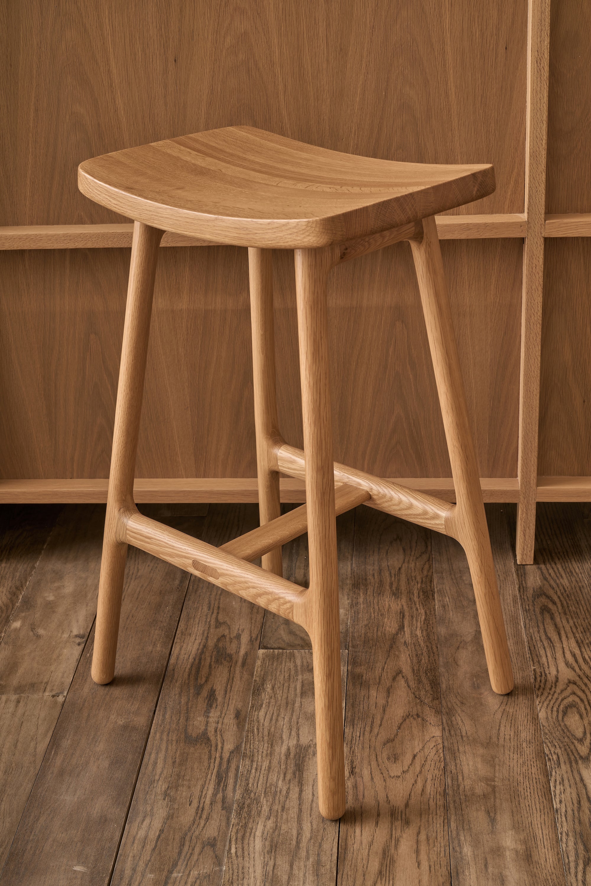 The Odd Counter Stool | Wood by Wooden Stories is a light solid oak, four-legged counter stool with a curved seat—an elegant piece of Danish design, shown here on a wooden floor before a wood-paneled wall.