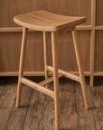 The Odd Counter Stool | Wood by Wooden Stories is a light solid oak, four-legged counter stool with a curved seat—an elegant piece of Danish design, shown here on a wooden floor before a wood-paneled wall.