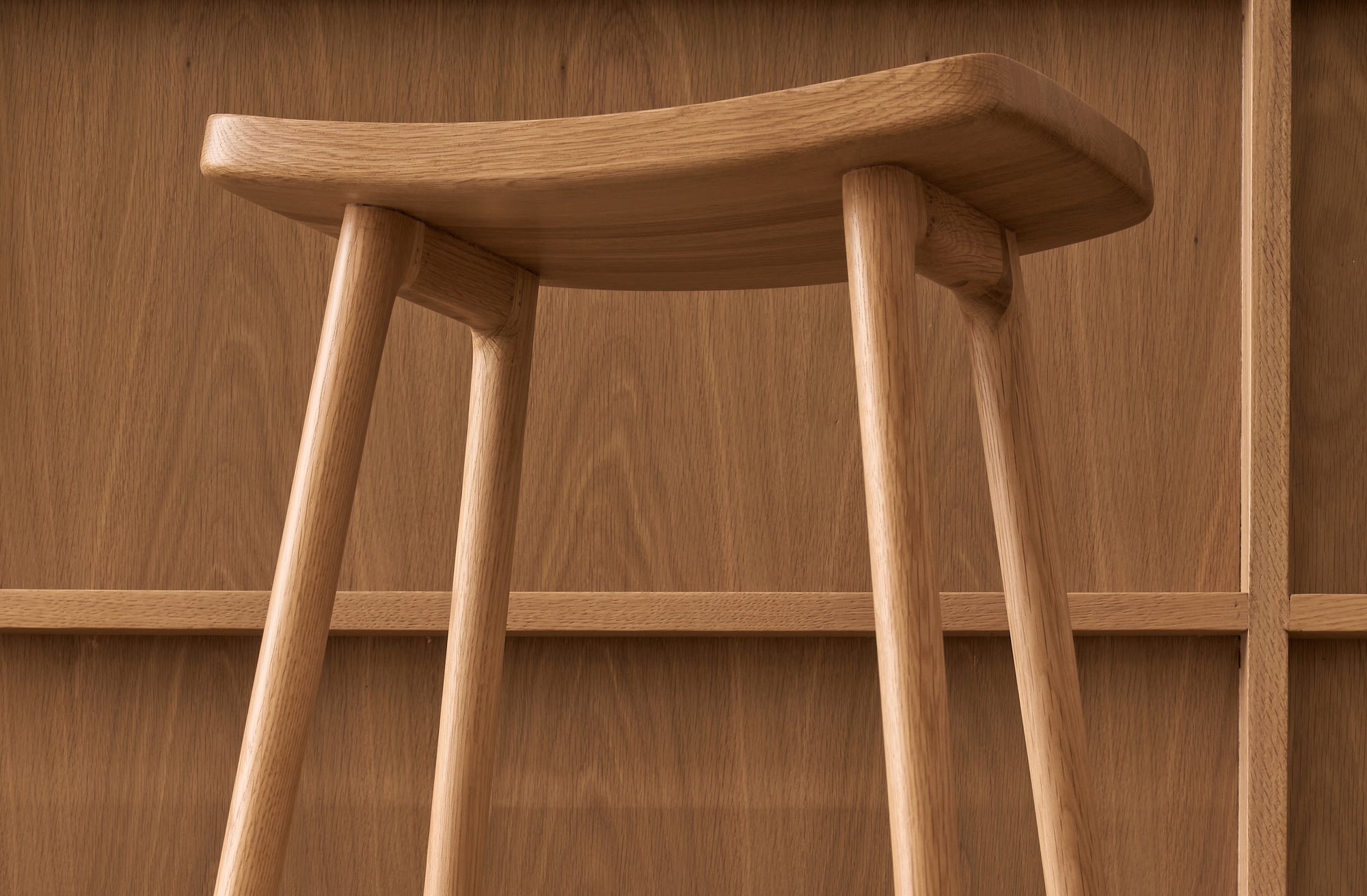 Close-up of the Odd Counter Stool | Wood by Wooden Stories, featuring four angled legs and a gently curved seat in solid oak, set before a matching wood-paneled wall—an elegant detail of Danish-inspired design.