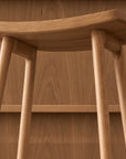 Close-up of the Odd Counter Stool | Wood by Wooden Stories, featuring four angled legs and a gently curved seat in solid oak, set before a matching wood-paneled wall—an elegant detail of Danish-inspired design.