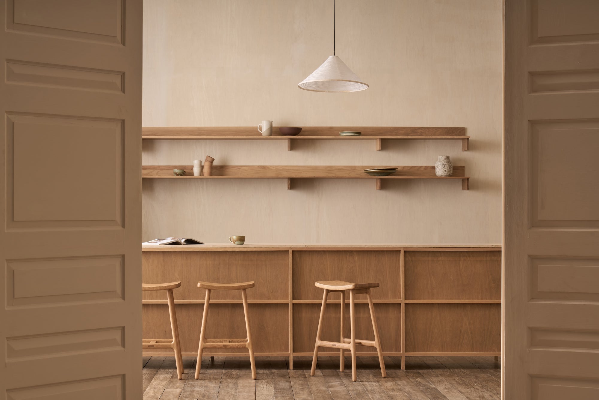 Minimalist kitchen featuring Wooden Stories' Odd Counter Stool | Wood, wooden shelves, Danish-inspired furniture, and simple ceramics. Neutral beige walls and soft natural light create a calm atmosphere. Scene viewed through open double doors.