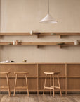 Minimalist kitchen featuring Wooden Stories' Odd Counter Stool | Wood, wooden shelves, Danish-inspired furniture, and simple ceramics. Neutral beige walls and soft natural light create a calm atmosphere. Scene viewed through open double doors.