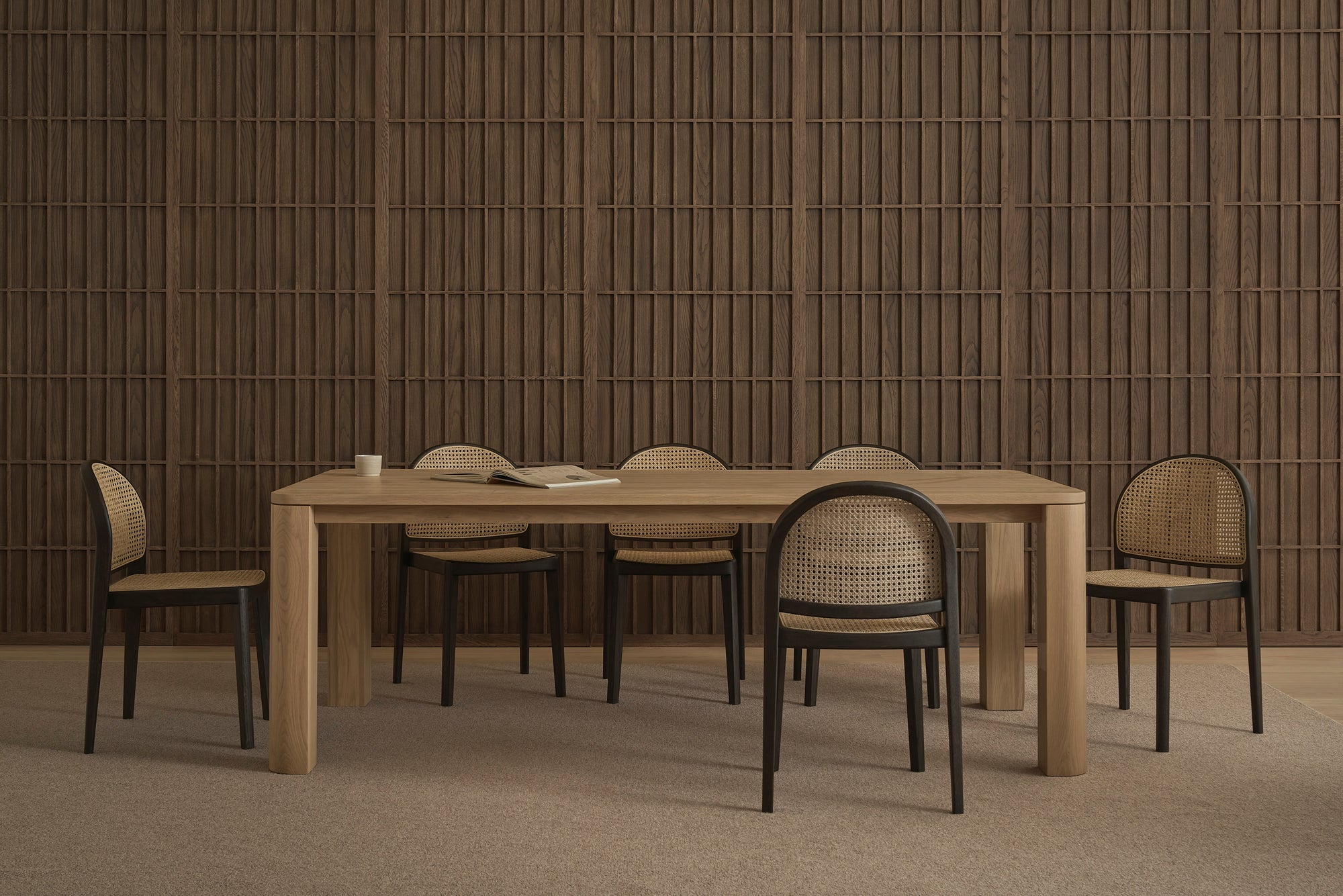 A minimalist dining room showcases a wooden table and six Wooden Stories Luk Dining Chairs with cane weave rattan backs, plus a cup and open book, set against a textured wooden wall and neutral carpeted floor.