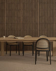 A minimalist dining room showcases a wooden table and six Wooden Stories Luk Dining Chairs with cane weave rattan backs, plus a cup and open book, set against a textured wooden wall and neutral carpeted floor.