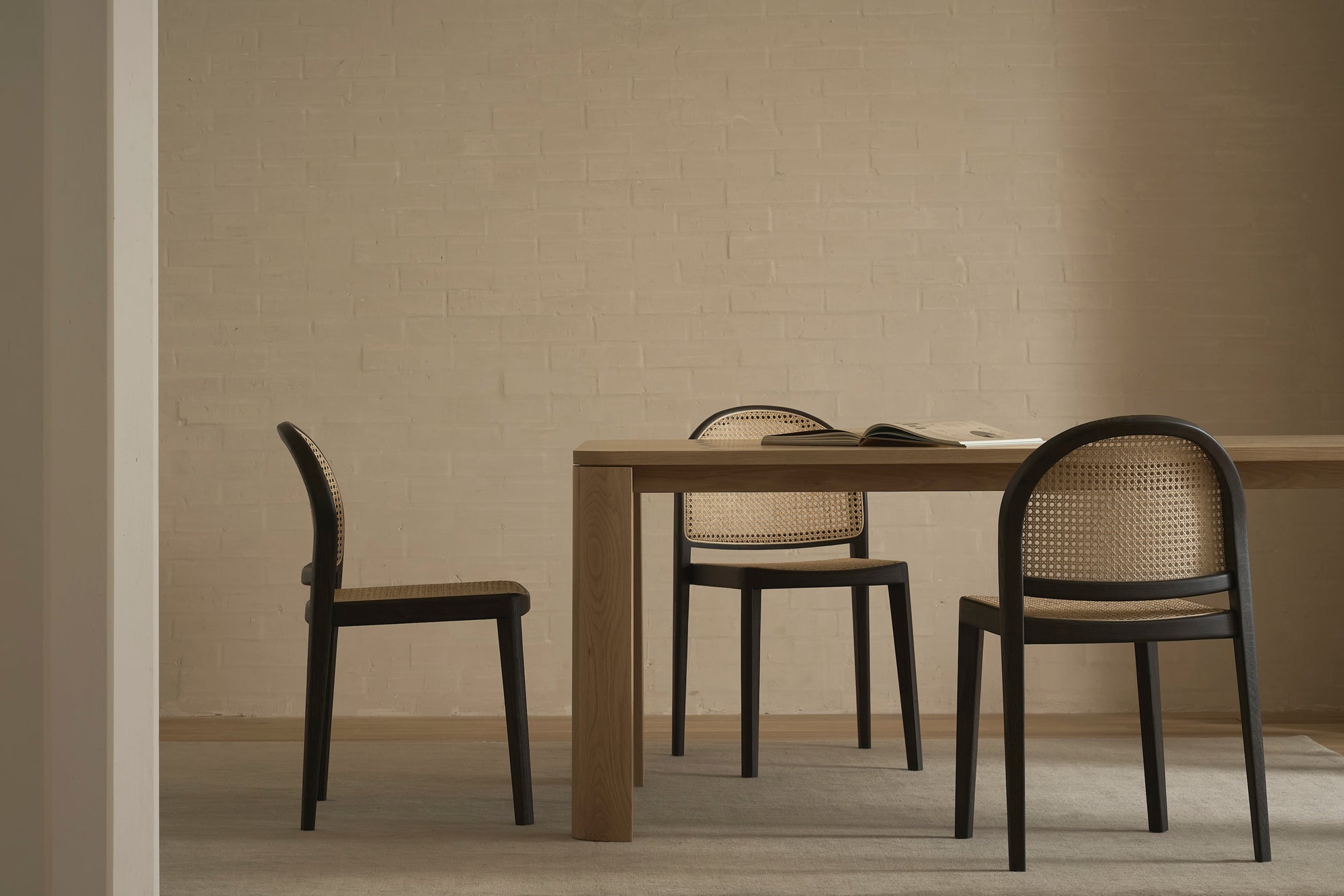 A minimalist dining area features the Wooden Stories Oli Dining Table and three woven cane chairs, set by a light beige brick wall. An open book rests on the table, completing this calm and neutral Danish-inspired setting.